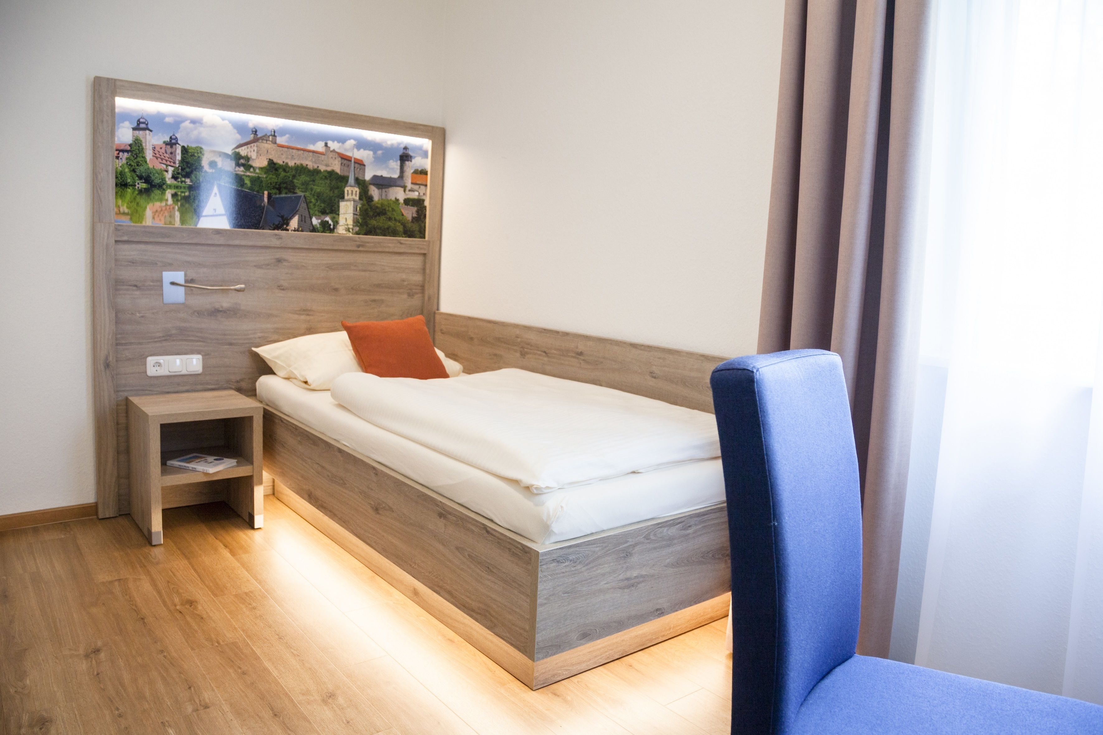 Ein modernes Hotelzimmer mit einem hölzernen Kopfteil, einem Einzelbett mit weißer Bettwäsche, einem kleinen Nachttisch mit einer Zeitschrift und Blick auf ein historisches Gebäude durch ein Fenster.