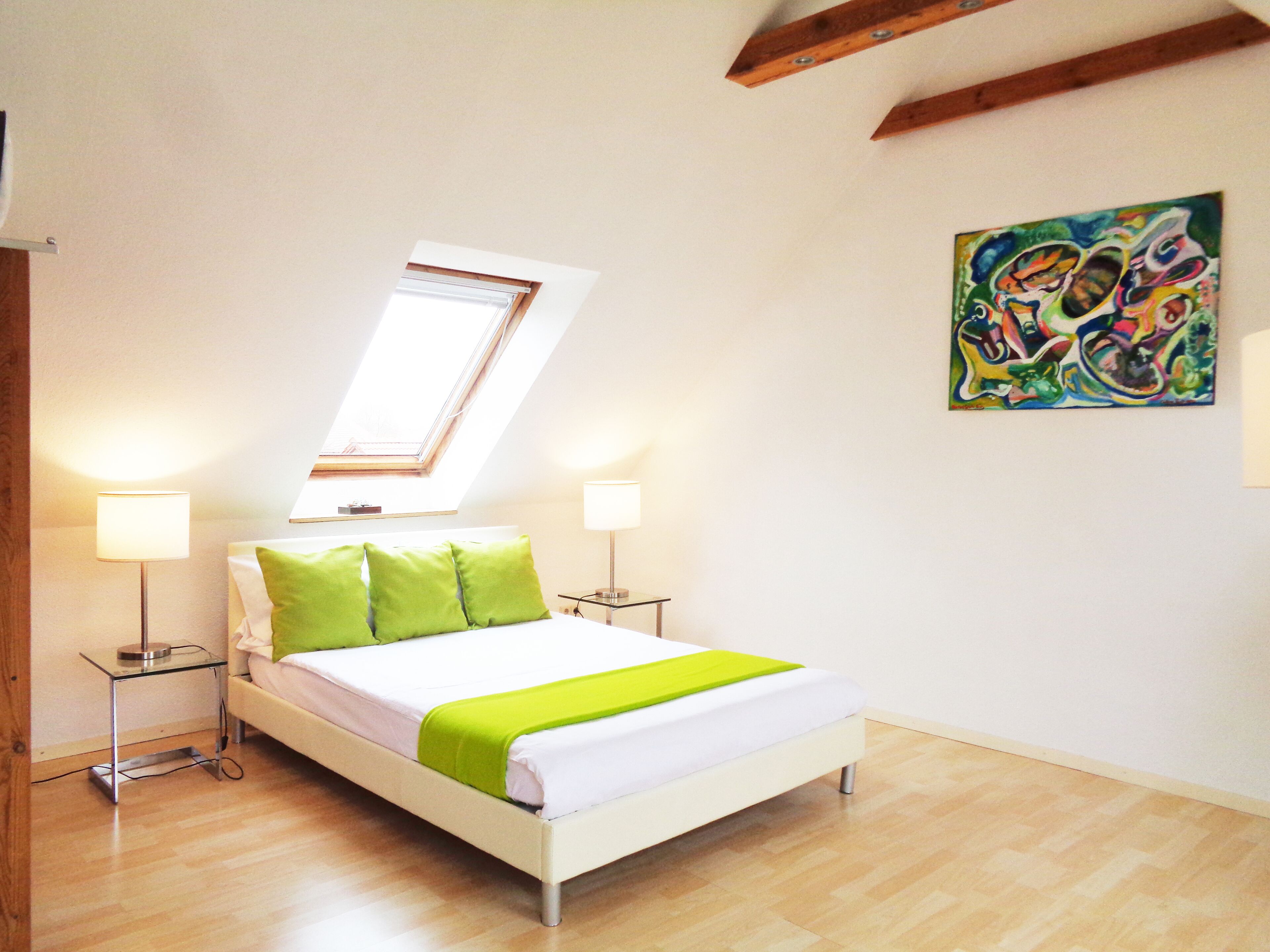 A bedroom with a skylight, a bed with green pillows, bedside lamps, and a colorful abstract painting on the wall.