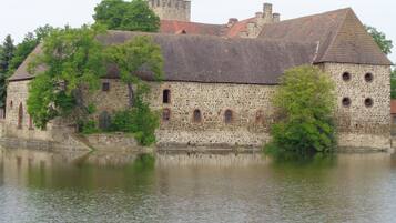 Un bâtiment historique en pierre, doté d’arcs en plein cintre au niveau des fenêtres et surmonté d’une tour, situé au bord d’une étendue d’eau paisible.