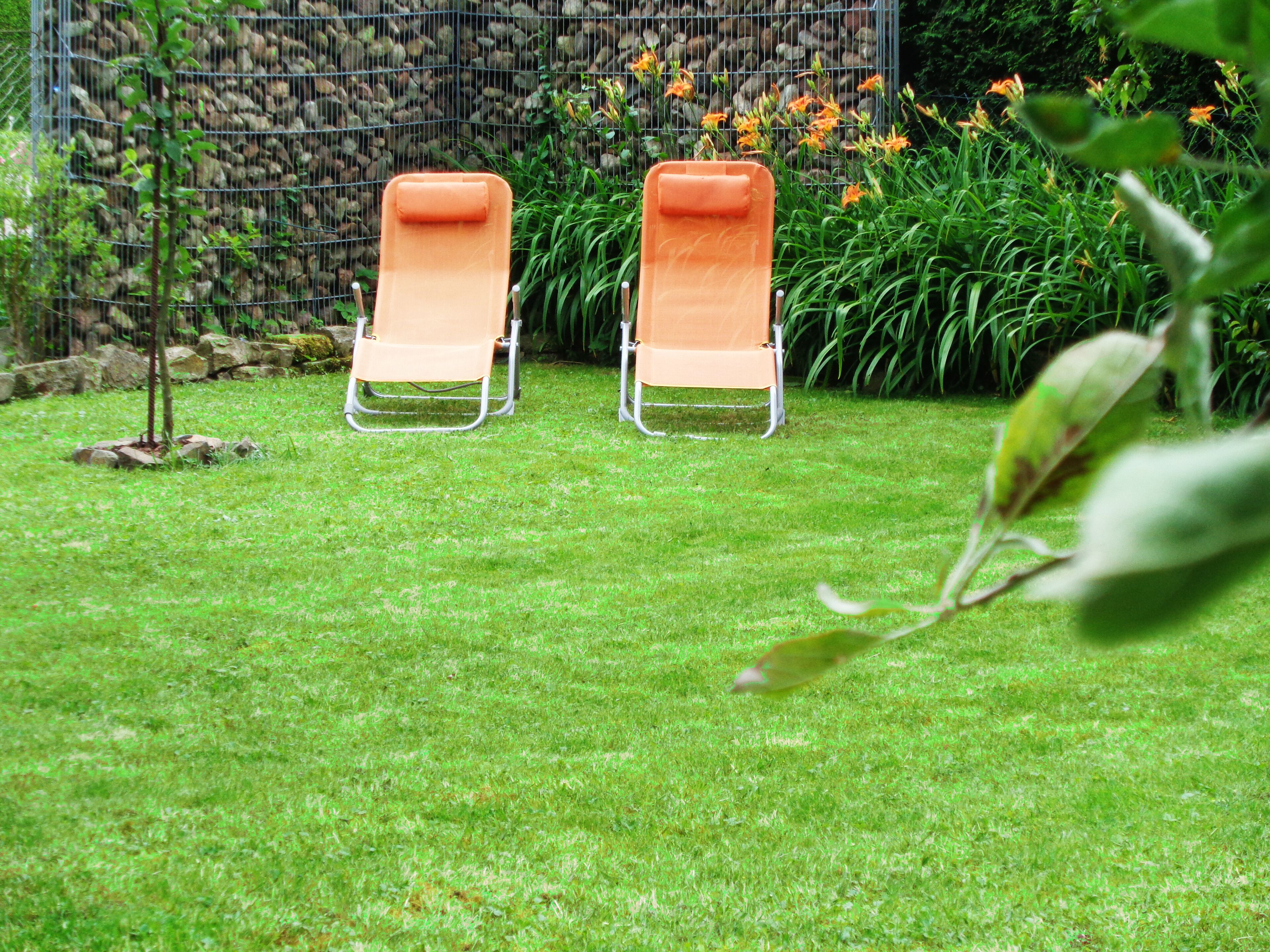 Dos sillones de exterior en un área verde, con un muro de piedra y plantas con flores al fondo.