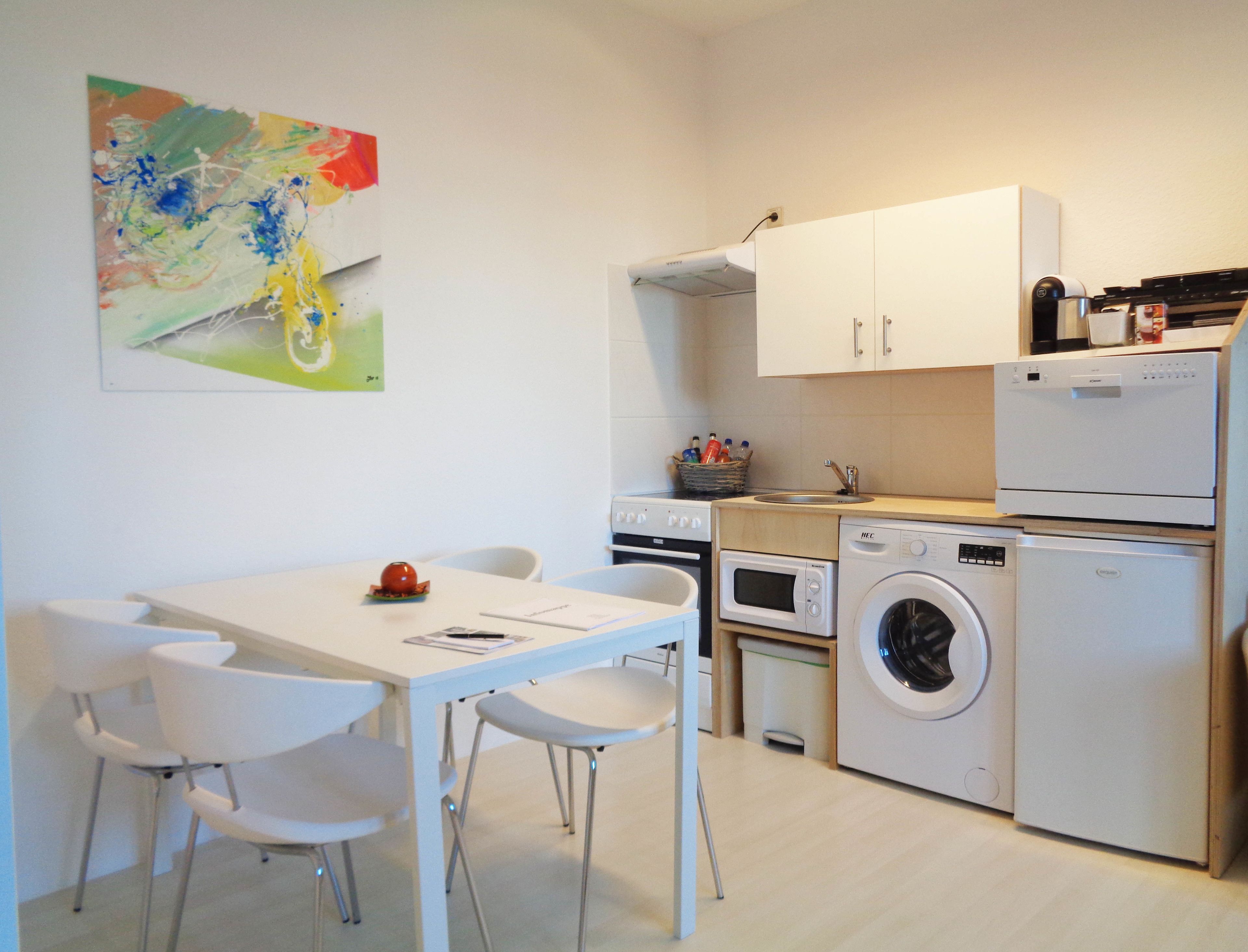 Une cuisine de taille réduite avec des appareils électroménagers blancs, une table à manger avec des chaises et un tableau abstrait aux couleurs vives accroché au mur.