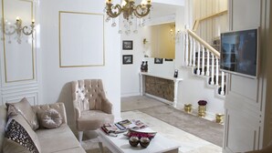 A living room with a beige sofa, a tufted armchair, a coffee table with magazines, a chandelier, and a fireplace. - Ozkavak Hotel Kaleici (Antalya)