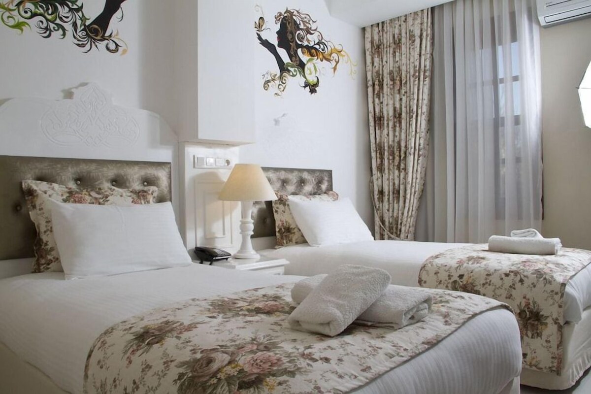 A hotel room with two beds, floral bedding, a decorative headboard, a bedside lamp, and a window with curtains.