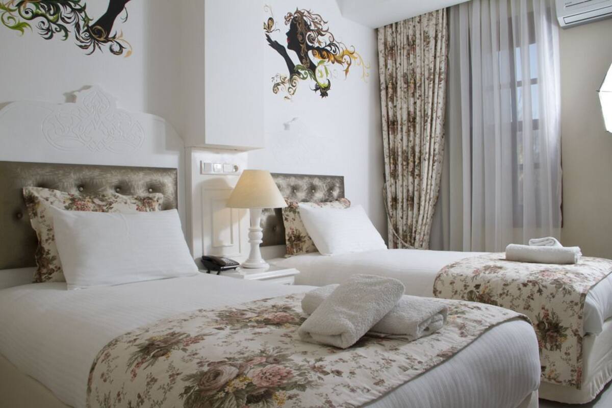 A hotel room with two beds, floral bedding, a decorative headboard, a bedside lamp, and a window with sheer curtains.