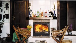 Una habitación acogedora con chimenea, dos sillas de madera, una mesita con un cuenco y una estantería con varias botellas.