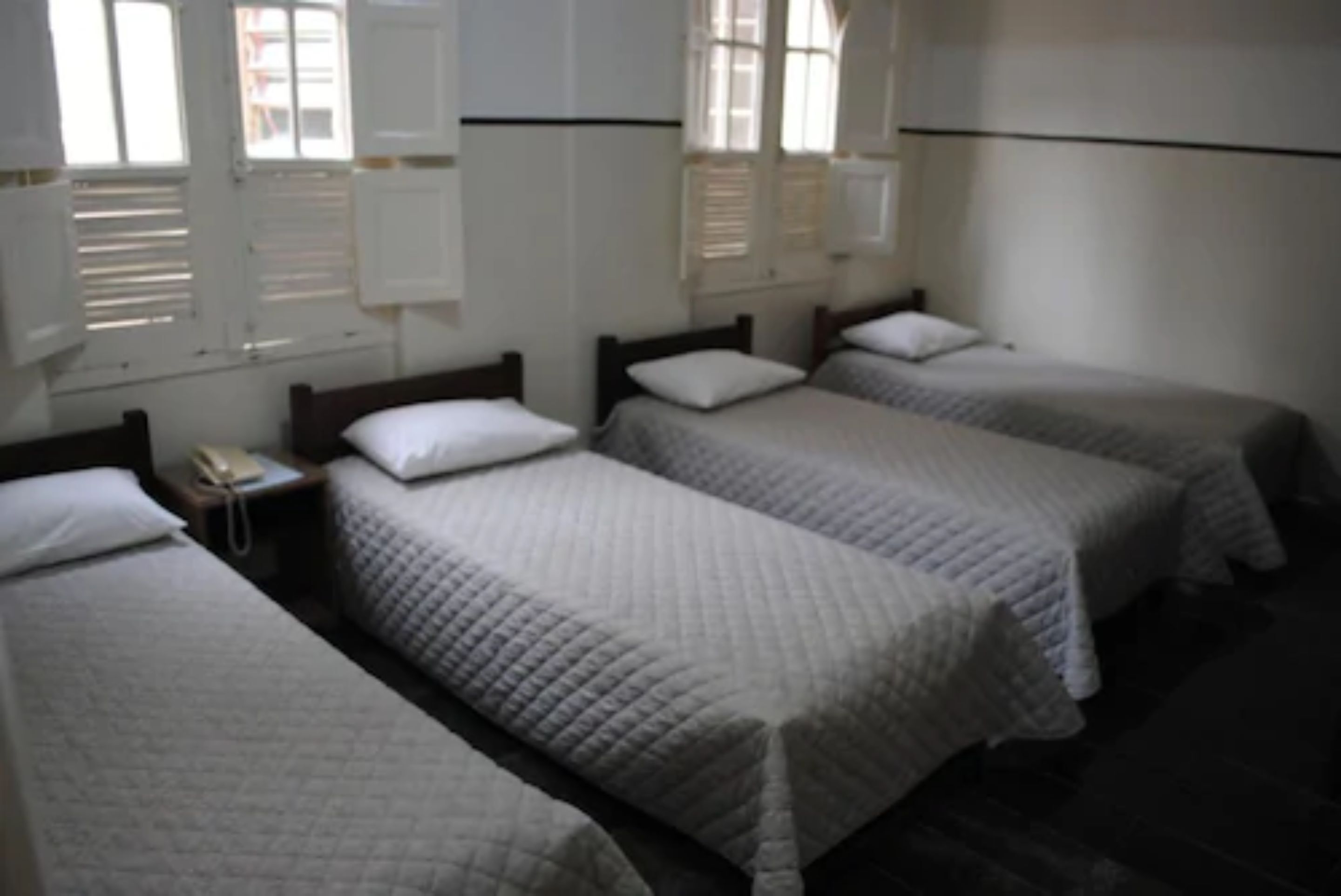 Quarto de hotel com três camas de solteiro, cada uma com travesseiro e cobertor. Há uma pequena mesa de cabeceira com uma luminária e um telefone.