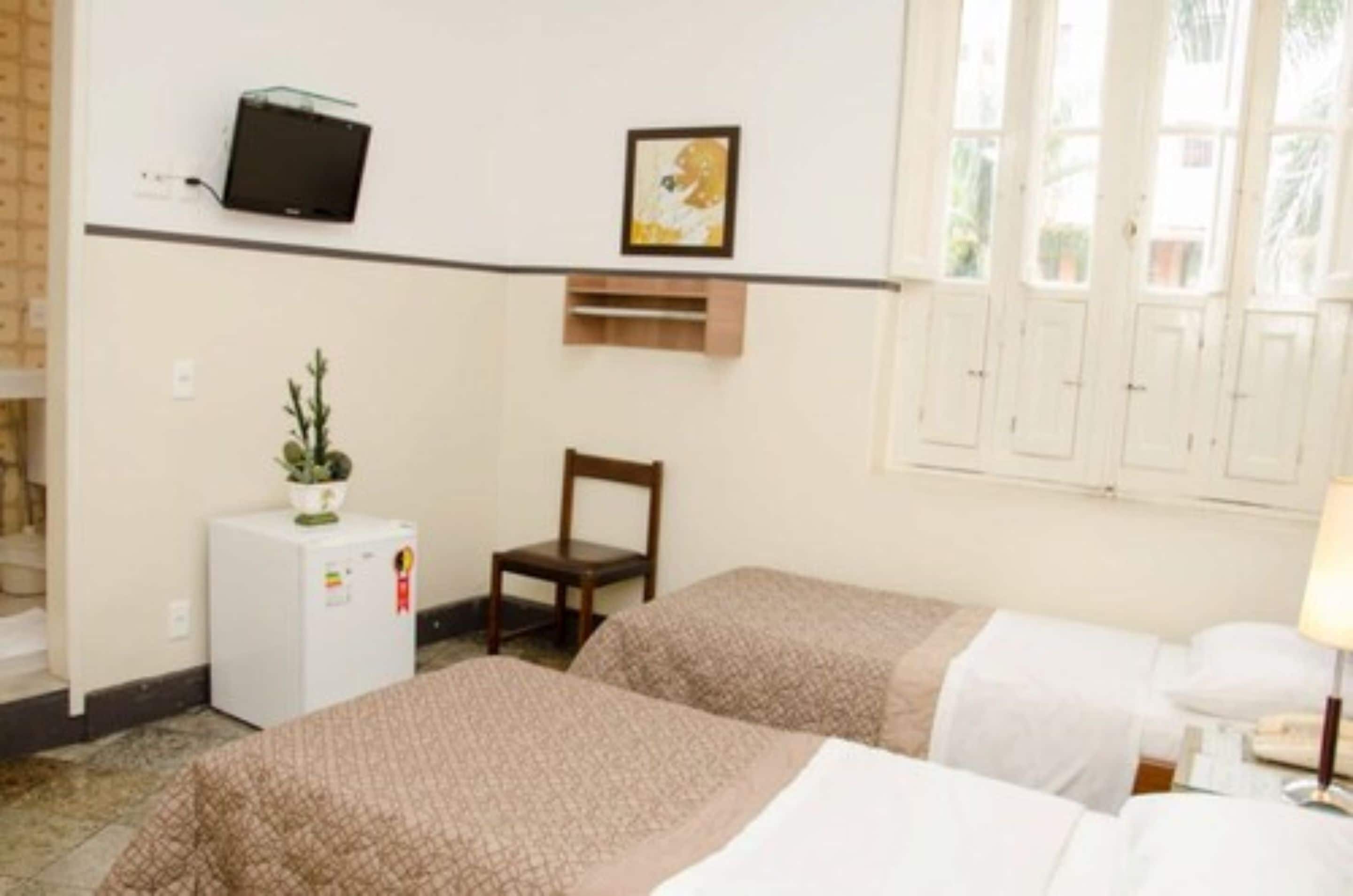 Una camera d'albergo con due letti, una piccola scrivania, una sedia, una televisione, un frigorifero e una finestra con tende.