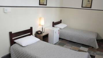 A hotel room with two single beds, each with a wooden headboard and a white pillow. There is a small bedside table with a lamp and a refrigerator. The floor has a patterned tile design.