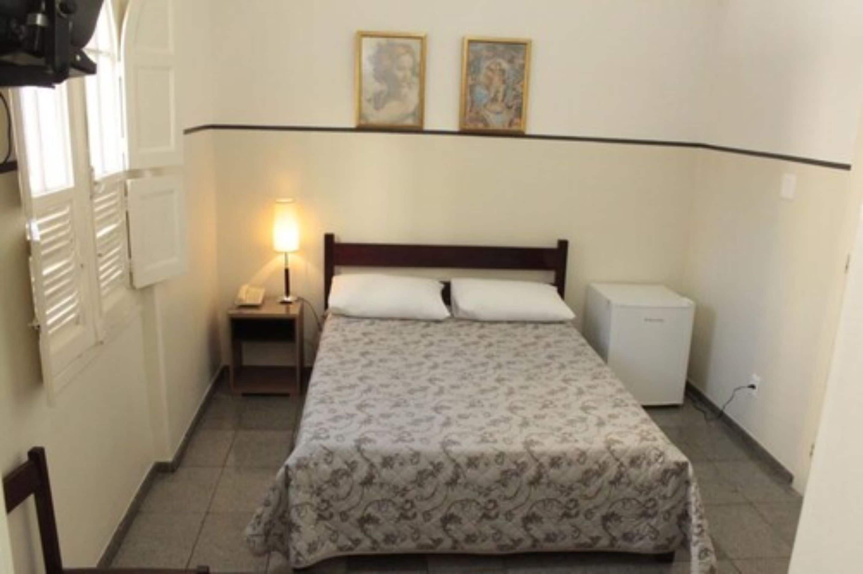 Una habitación pequeña y sencilla con cama, mesita de noche, lámpara y refrigerador.