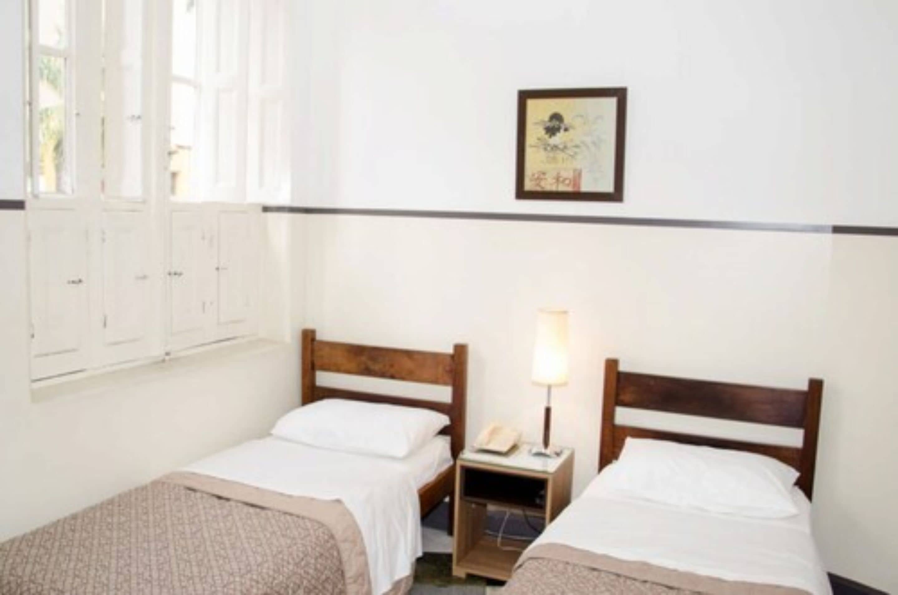 A room with two single beds, a nightstand with a lamp, a framed picture on the wall, and a window with shutters.