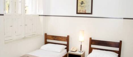 A room with two single beds, a nightstand with a lamp, a framed picture on the wall, and a window with shutters.