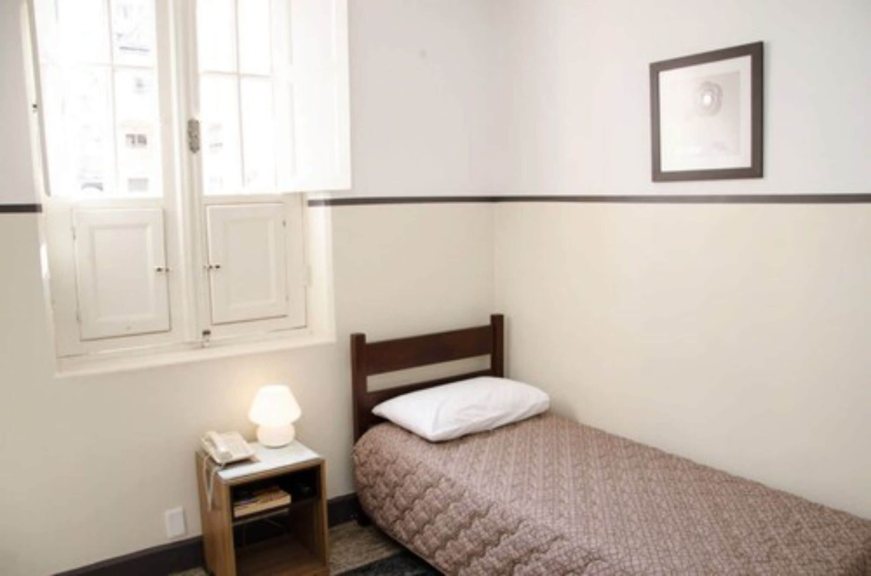 Quarto pequeno com uma cama de solteiro, cabeceira de madeira, luminária de cabeceira e um quadro na parede.