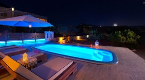 A swimming pool with a blue-lit edge, surrounded by lounge chairs and a large umbrella. - Melfe Villas (Rhodes)