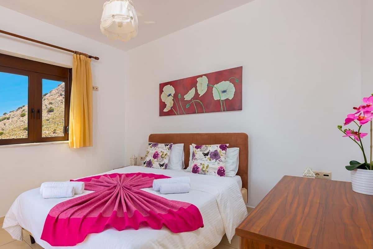 A bedroom with a bed, a wooden headboard, a window with curtains, a painting on the wall, and a table with a potted orchid.