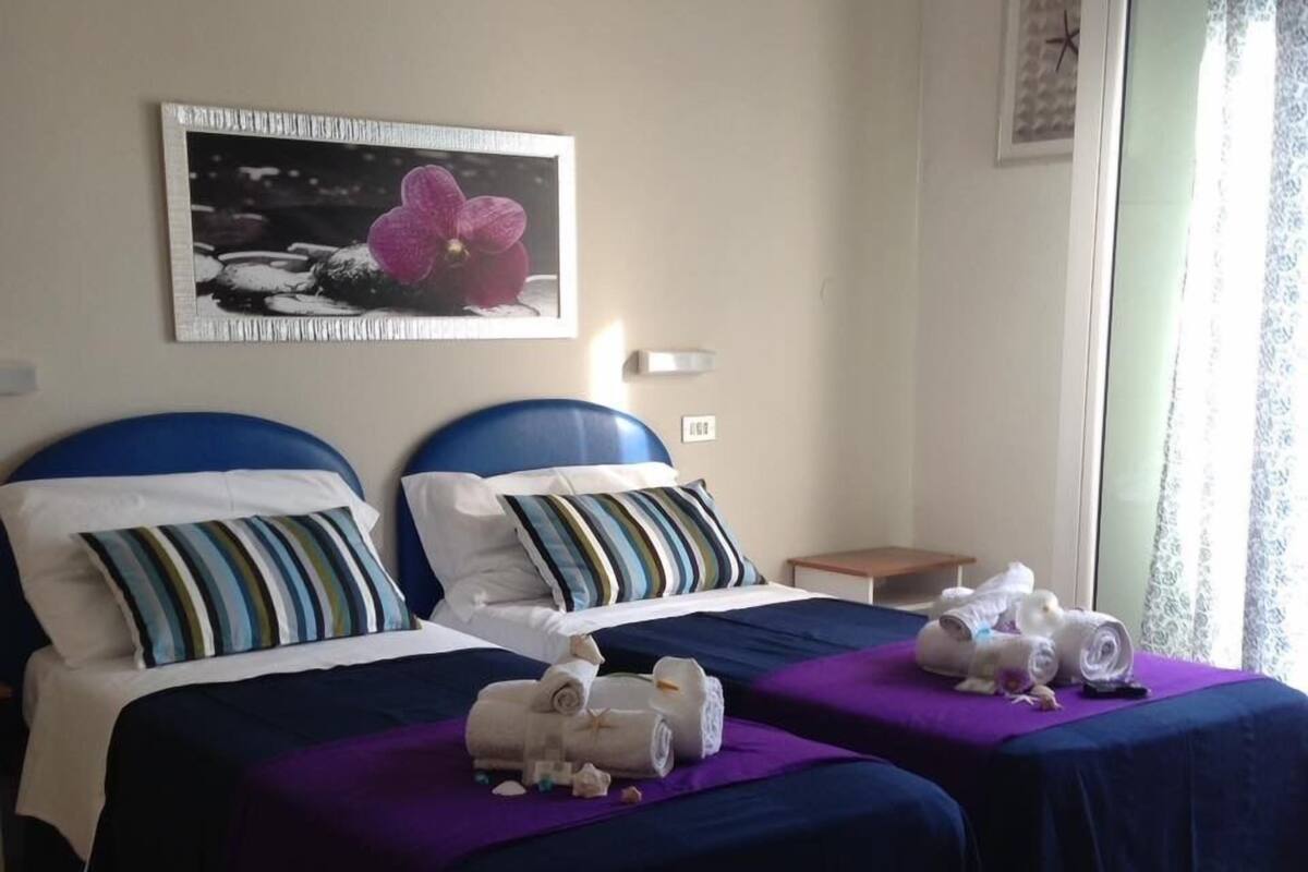 A hotel room with two beds, each with a blue headboard and striped pillows. There is a painting on the wall and a small table with a lamp.