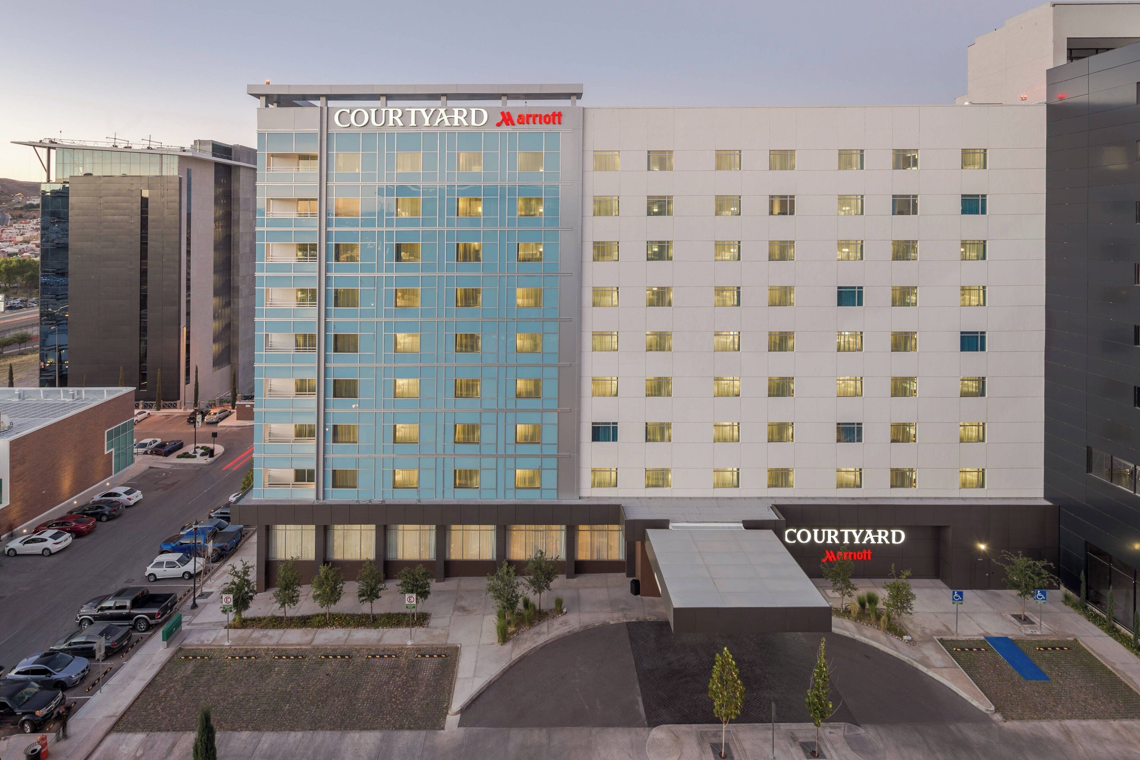 A Courtyard Marriott hotel with a modern design, featuring a mix of blue and white facades, a parking lot, and a landscaped area with trees.