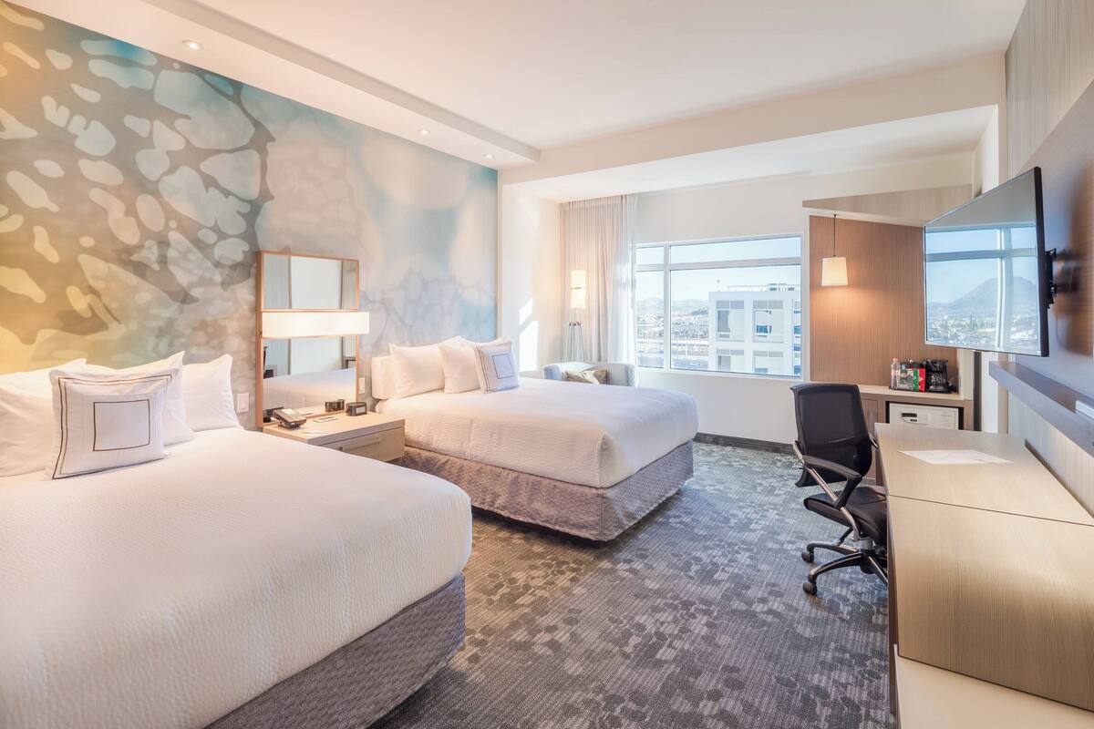 Habitación de hotel con dos camas, un escritorio, una silla, un televisor y un ventanal con vistas a la ciudad.
