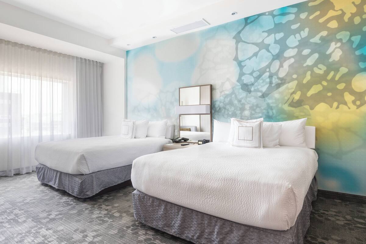 Habitación de hotel con dos camas, un ventanal con cortinas translúcidas y un mural abstracto de colores.