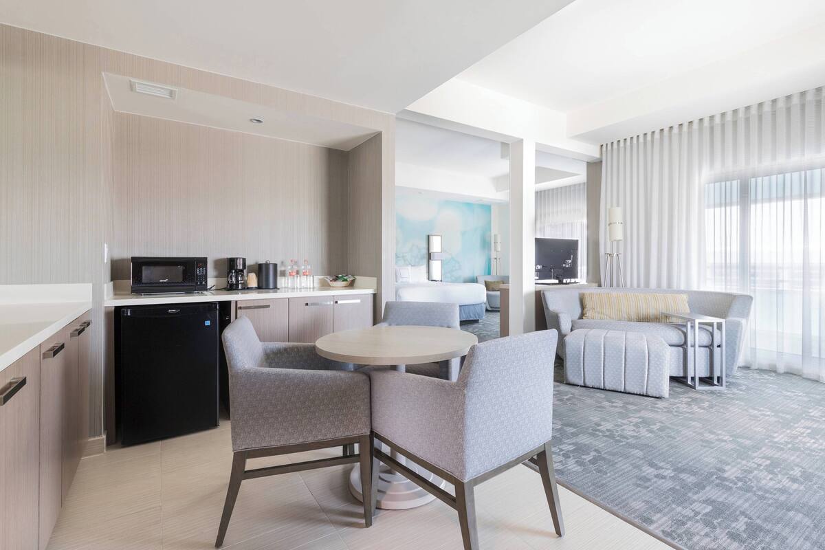 Una habitación de hotel moderna con kitchenette, un área de comedor con una mesa redonda y sillas, un sofá y un gran ventanal con cortinas.