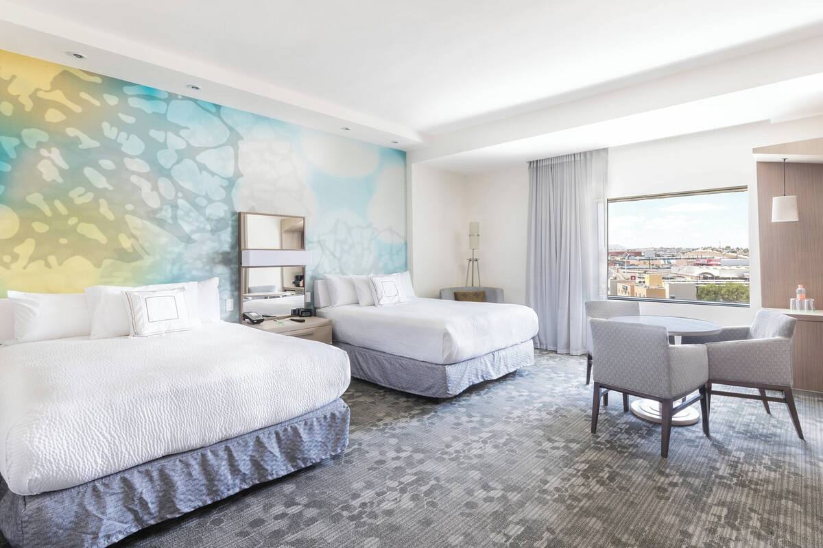 Habitación de hotel con dos camas, un escritorio, una silla y un amplio ventanal con vistas a la ciudad.
