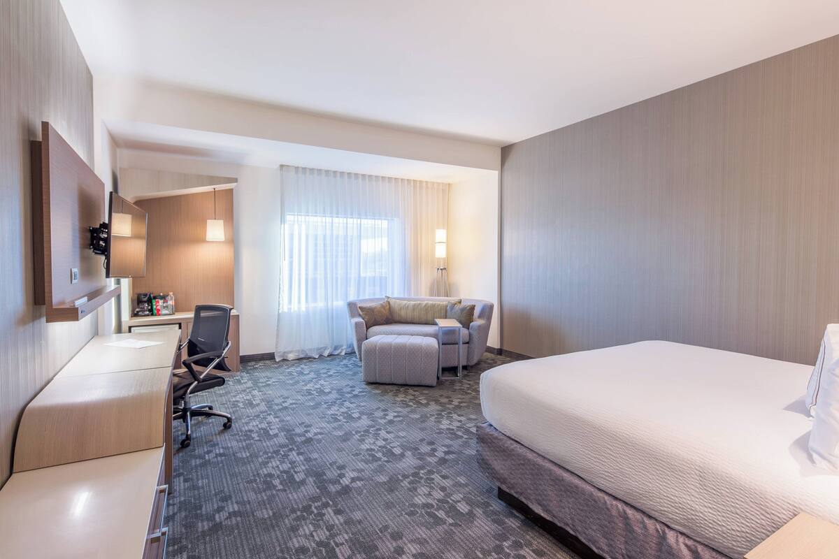 Una habitación de hotel con cama, escritorio, silla, sofá y un amplio ventanal con cortinas.