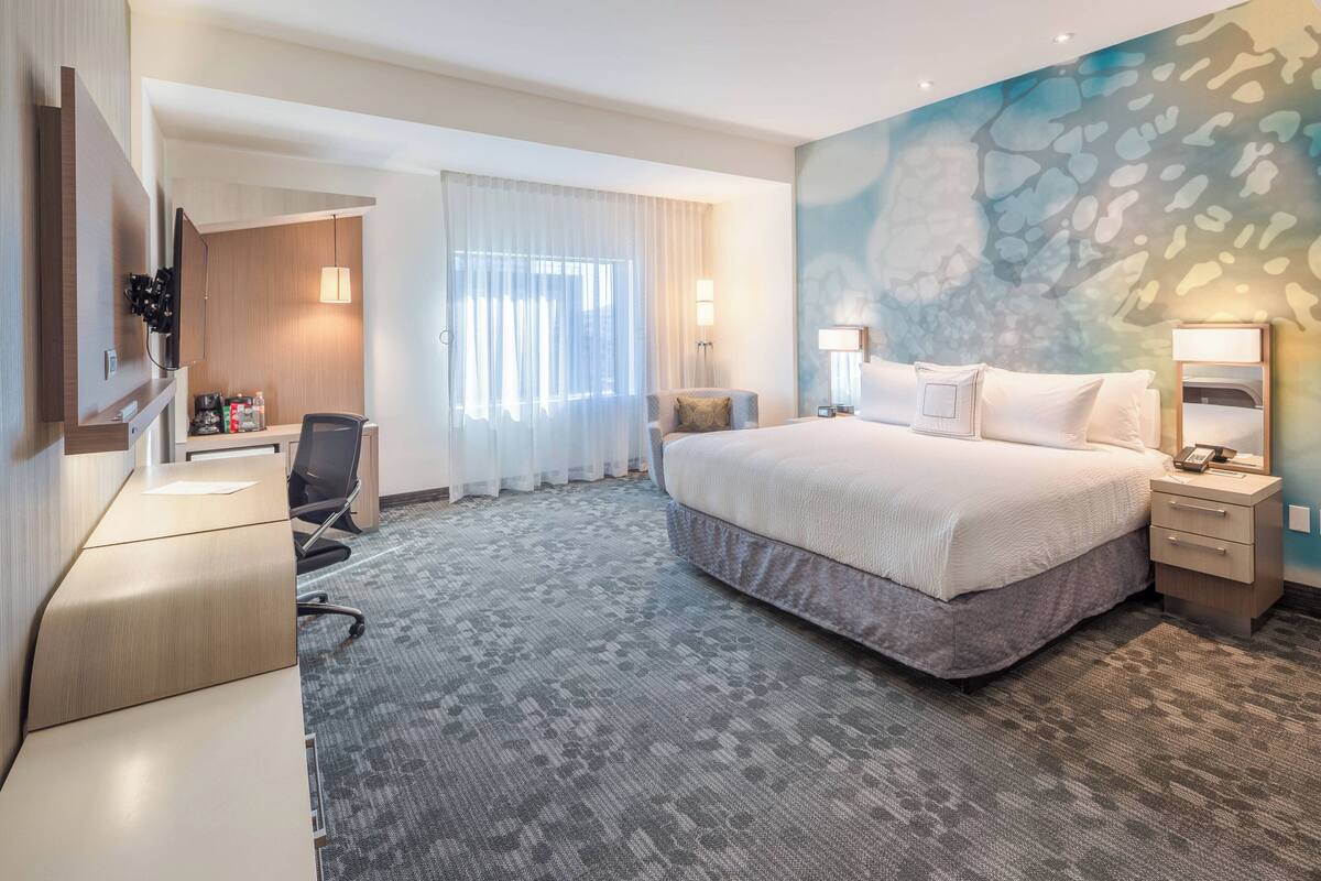 Habitación de hotel con cama, escritorio, silla y un ventanal con cortinas.