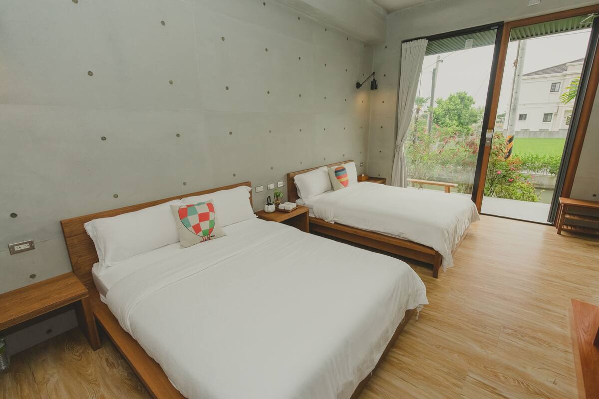 A hotel room with two beds, wooden headboards, white bedding, a small bedside table with a lamp, and a view of greenery outside.