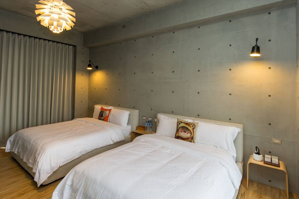 A hotel room with two beds, a wooden floor, a concrete accent wall, and a ceiling light fixture.