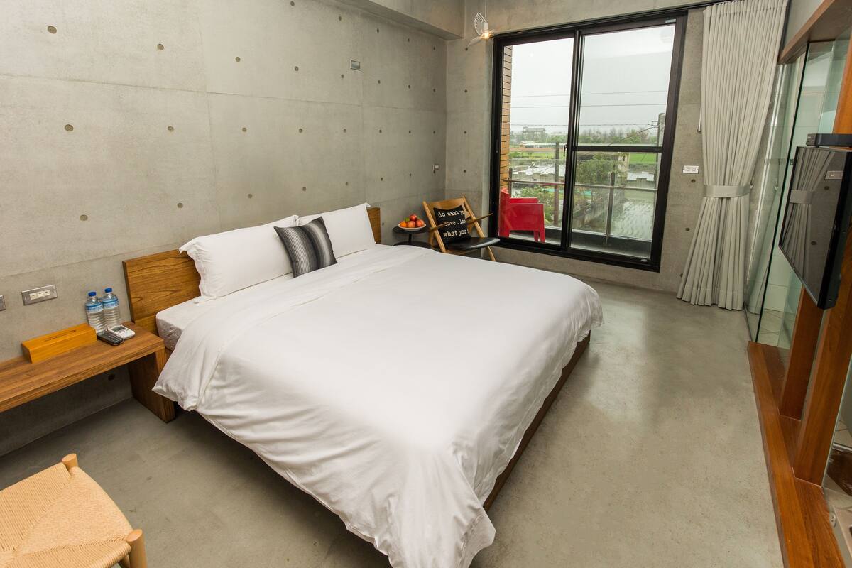 A hotel room with a large bed, a wooden headboard, a small table with a chair, a TV, and a window with curtains.