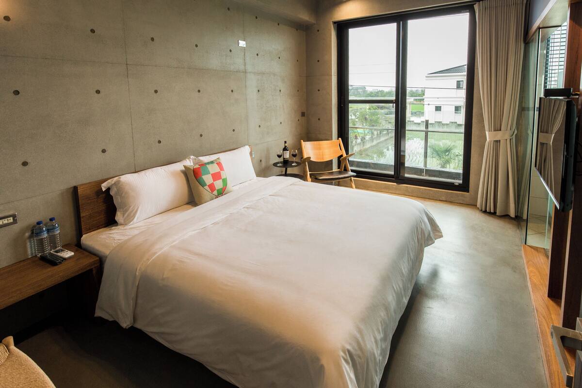 A hotel room with a large bed, a nightstand with a remote control and water bottles, a chair by the window, and a view of the outside.