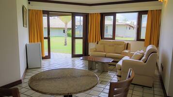 A living room with a sofa, a coffee table, and a dining table. There is a refrigerator and a window with curtains.