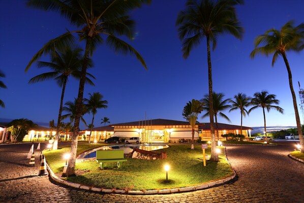 Un hotel con camino de entrada pavimentado, palmeras y una entrada bien iluminada.