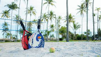 A beach volleyball court with a net, two volleyballs, and palm trees in the background.