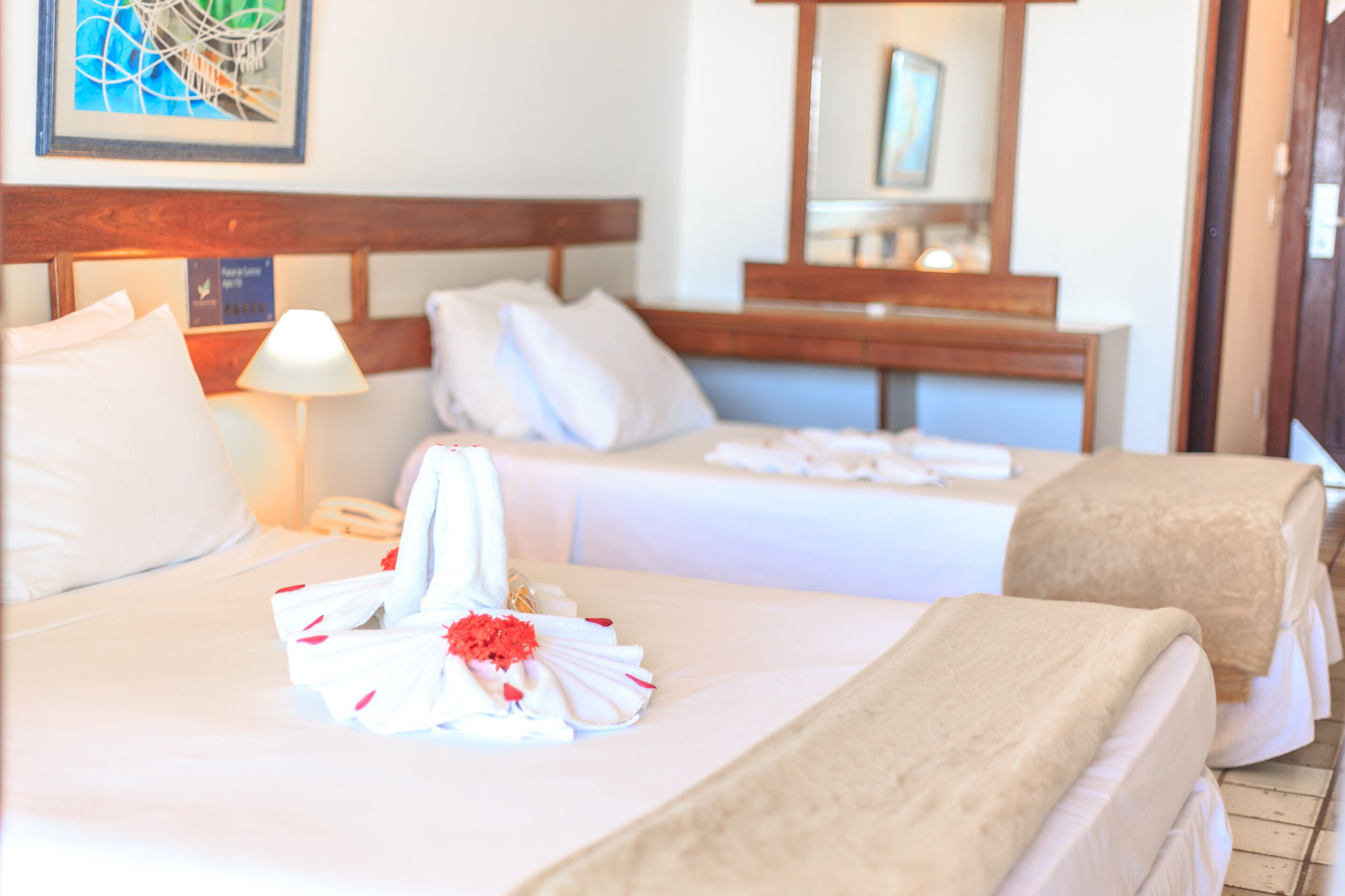 A hotel room with two beds, each with white linens and pillows, a bedside table with a lamp, and a framed artwork on the wall.