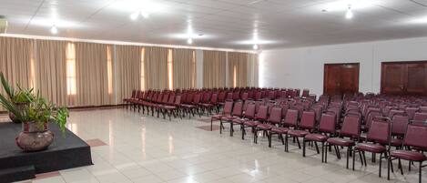 A large hall with rows of red chairs, a stage with a potted plant, and a tiled floor.