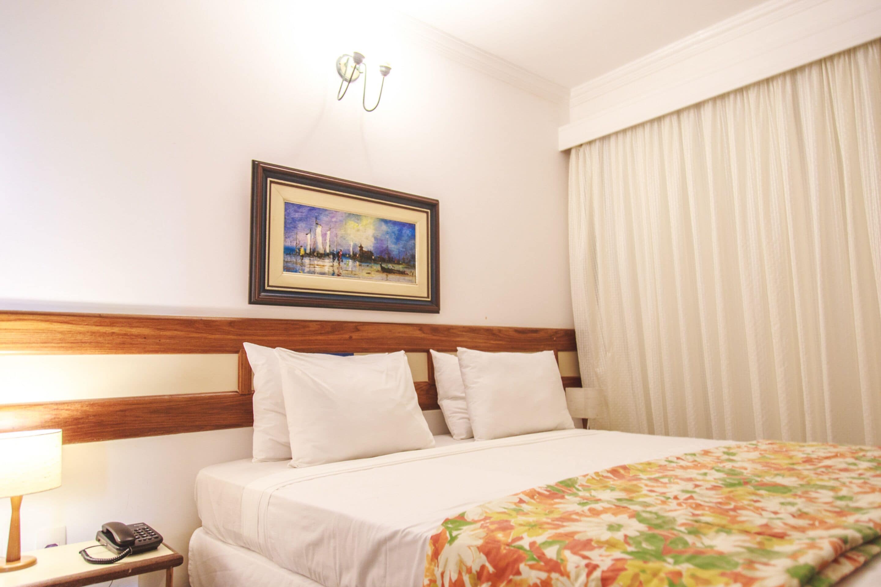 A hotel room with a bed, a nightstand with a phone, a painting on the wall, and a window with curtains.
