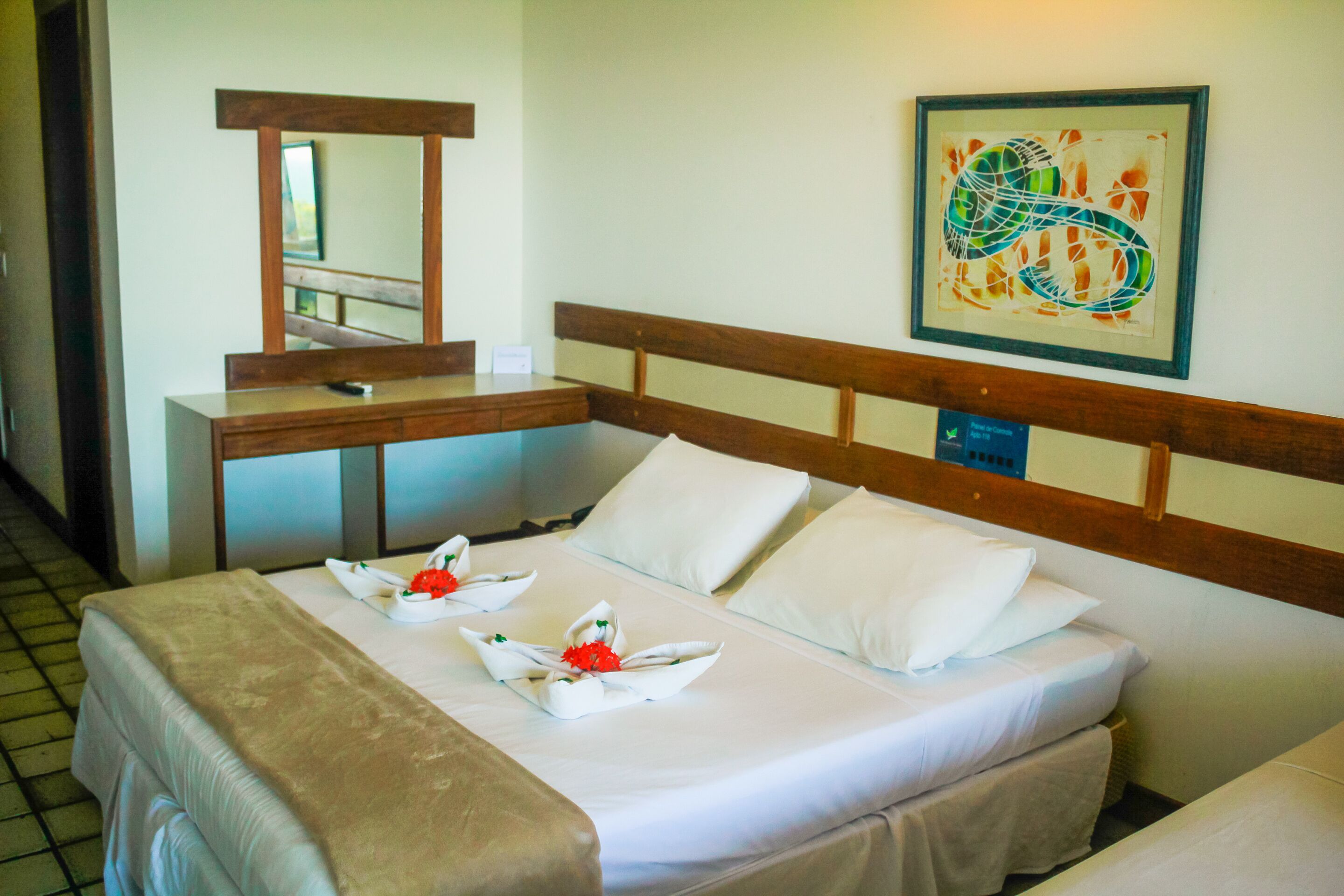 A hotel room with a neatly made bed, a wooden headboard, a mirror, and a framed abstract painting on the wall.
