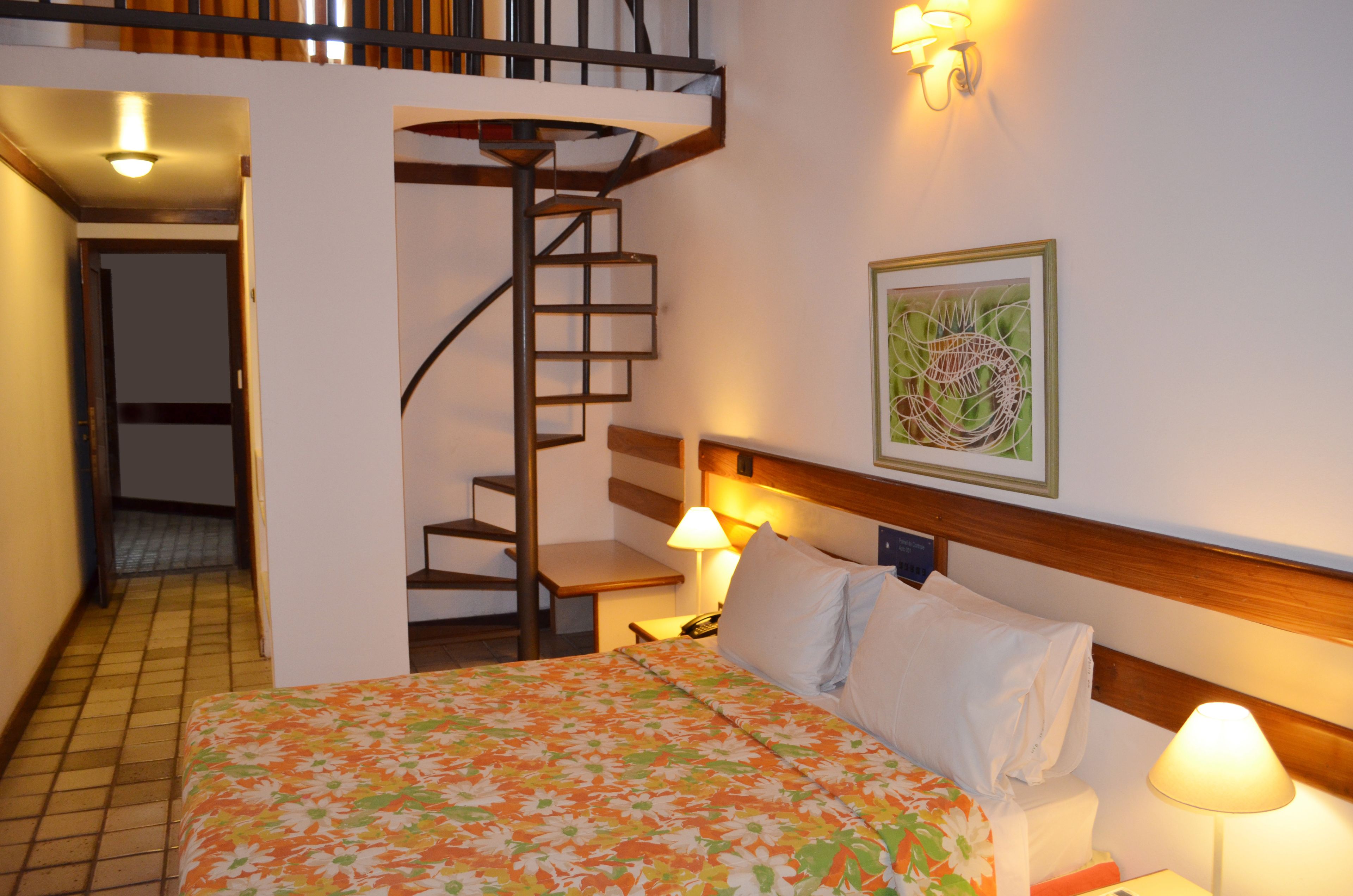 A hotel room with a bed, a staircase, a wall-mounted lamp, and a framed picture.