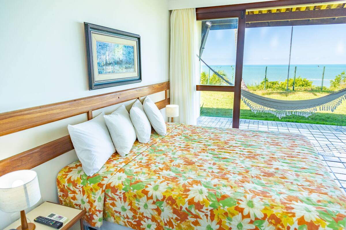 Ein Hotelzimmer mit einem Bett, einem Nachttisch, einer Lampe, einem gerahmten Bild und Blick auf das Meer durch eine Glastür.