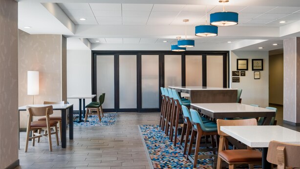 A modern dining area with multiple tables and chairs, a patterned rug, and blue pendant lights.