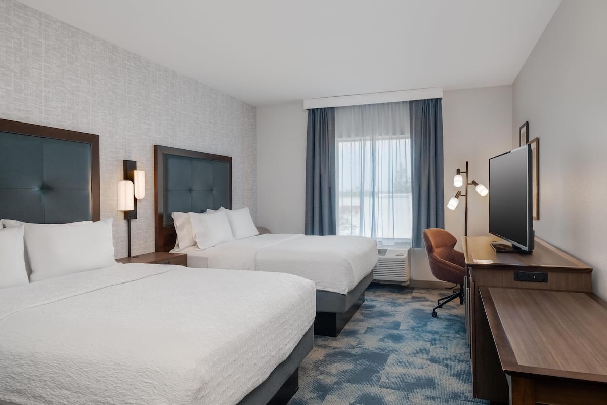 A hotel room with two beds, a desk with a TV, a chair, and a large window with curtains.