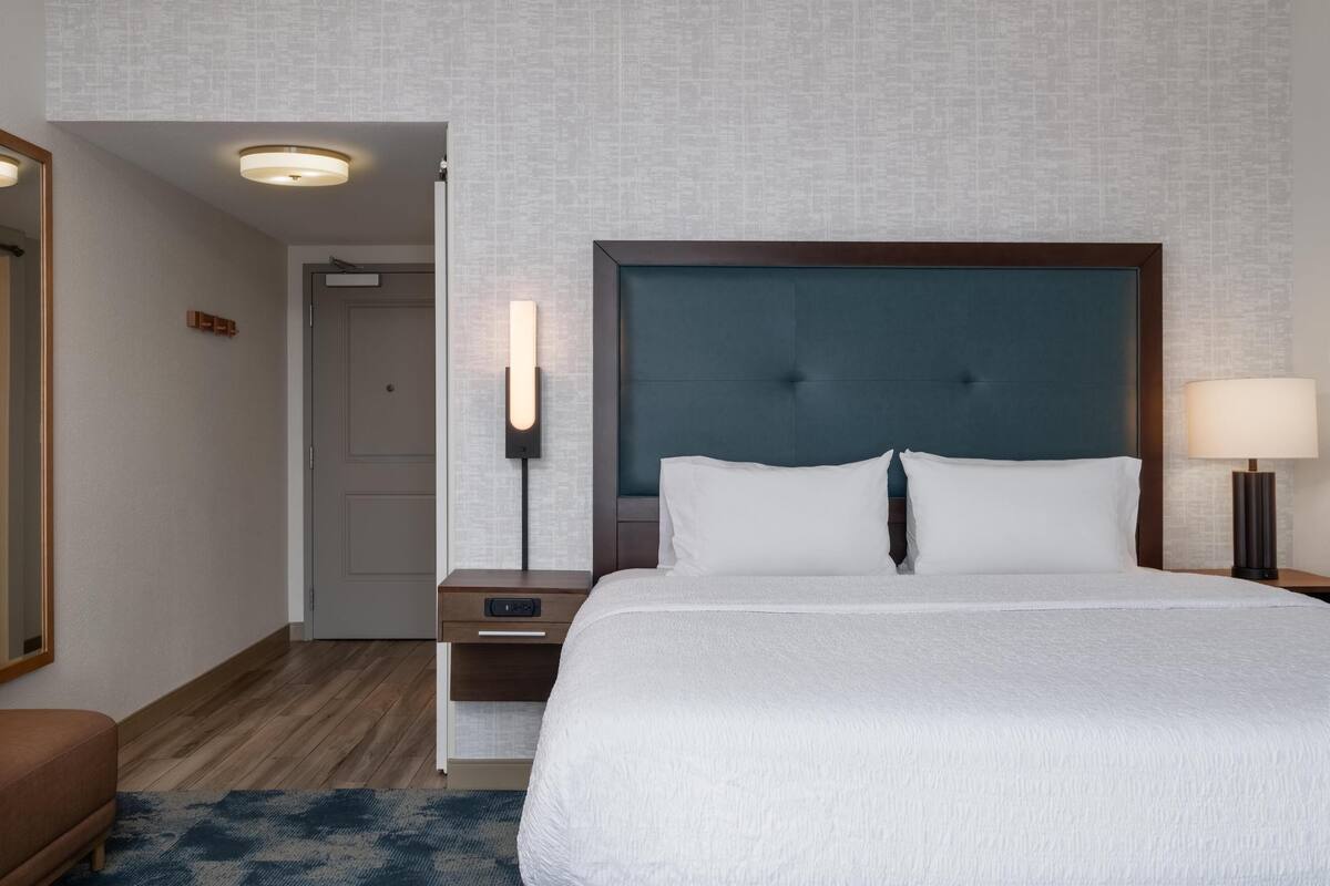 A hotel room with a large bed, a bedside table, a nightstand, a wall-mounted lamp, and a door leading to another room.