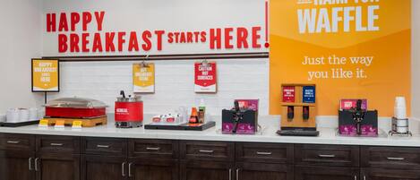 A breakfast station with a sign that reads "HAPPY BREAKFAST STARTS HERE!" and a sign for "THE HAMPTON WAFFLE" with a tagline "Just the way you like it."