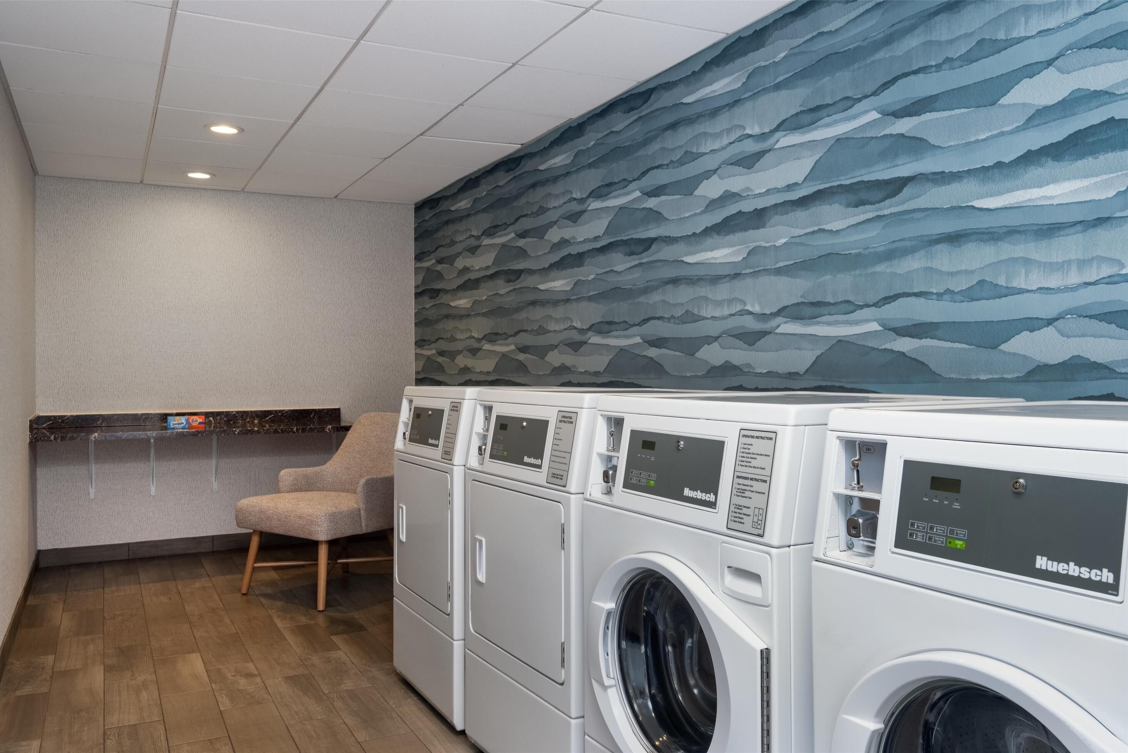 Een wasserette met een rij witte Huebsch wasmachines, een grijze stoel en een muur met een blauw bergdecor.