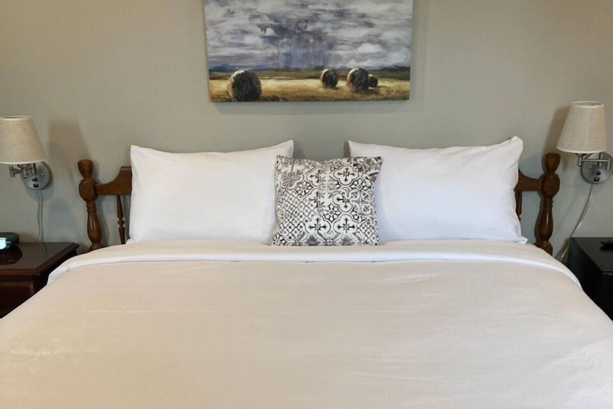 A neatly made bed with white linens, a patterned pillow, and a painting of hay bales on the wall above.