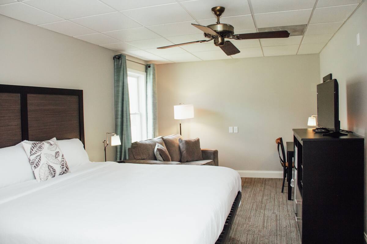 A hotel room with a large bed, a desk, a chair, a television, and a ceiling fan.