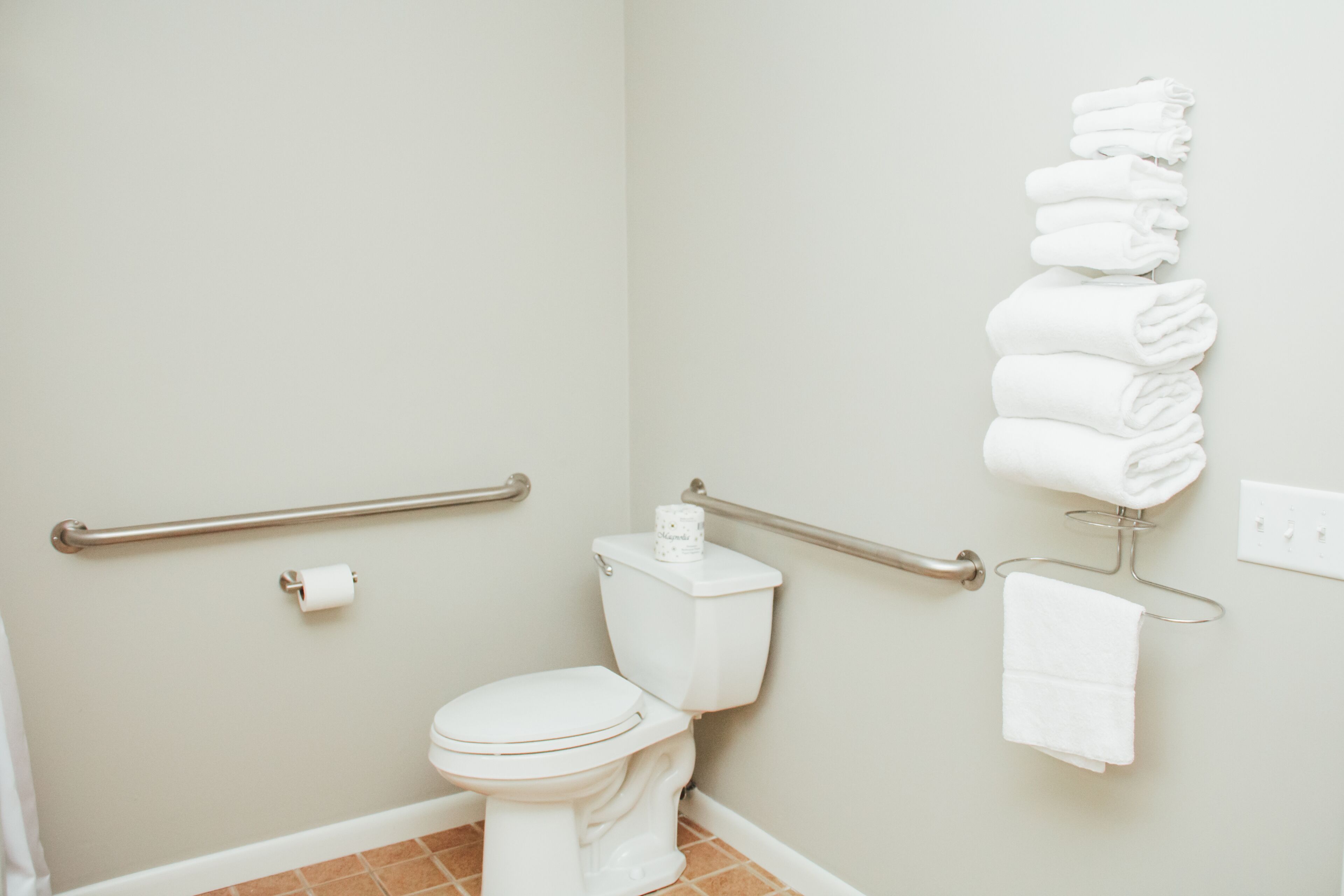 A bathroom with a toilet, a towel rack with folded towels, a handrail, and a toilet paper roll.