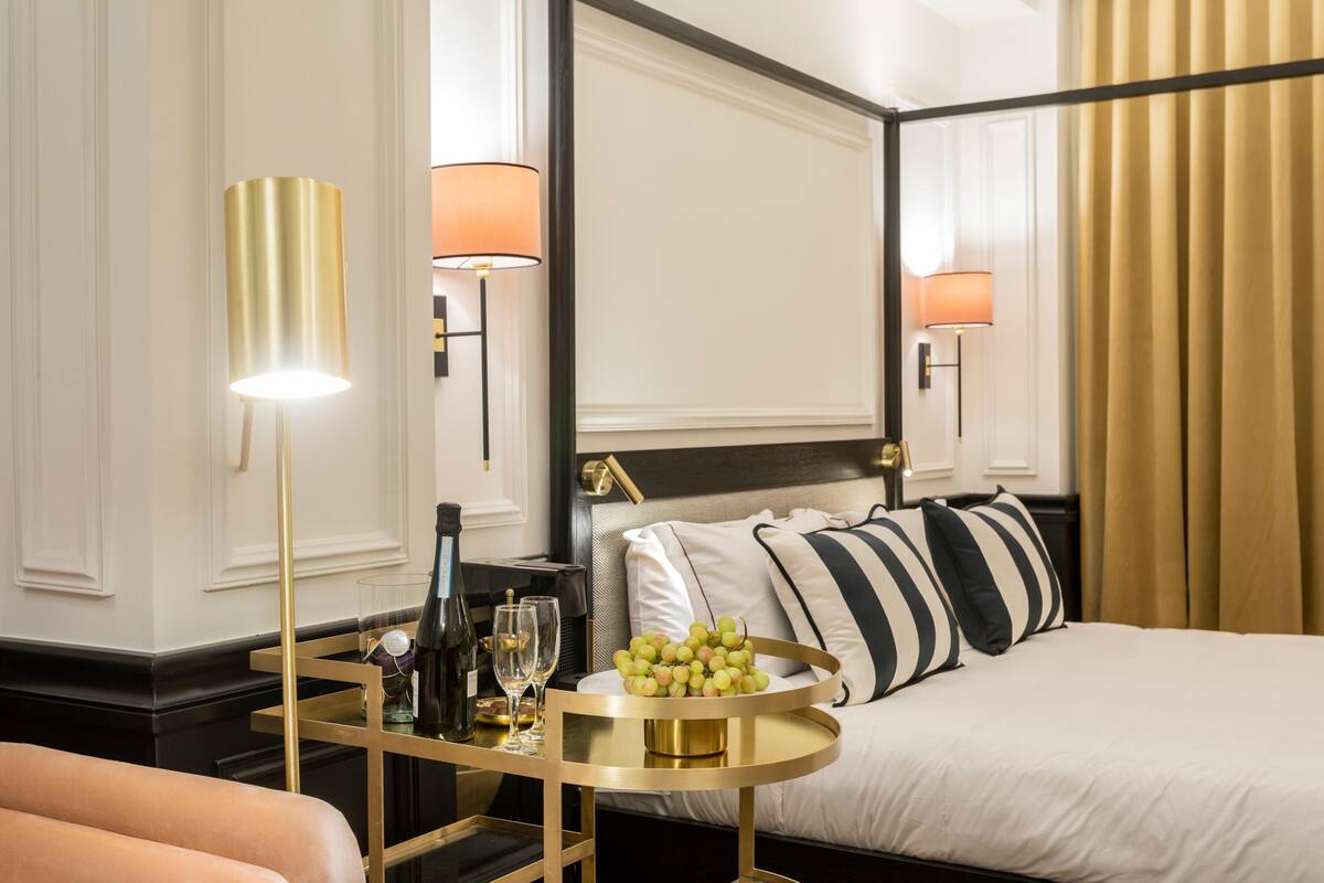 A hotel room with a bed, bedside lamps, a round table with a tray of fruit, and a minibar.