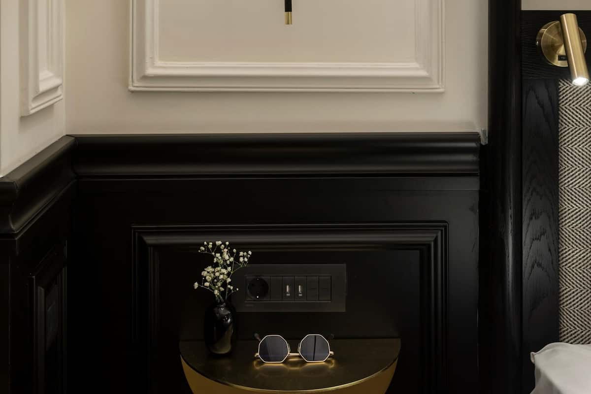 A wall-mounted lamp with a cylindrical shade, a black switch plate, and a small table with a vase and a pair of glasses.