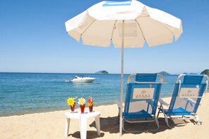 A beach scene with a white umbrella, two blue beach chairs, and a table with drinks and sunshades. - Atena Praia Hotel (São Sebastião)